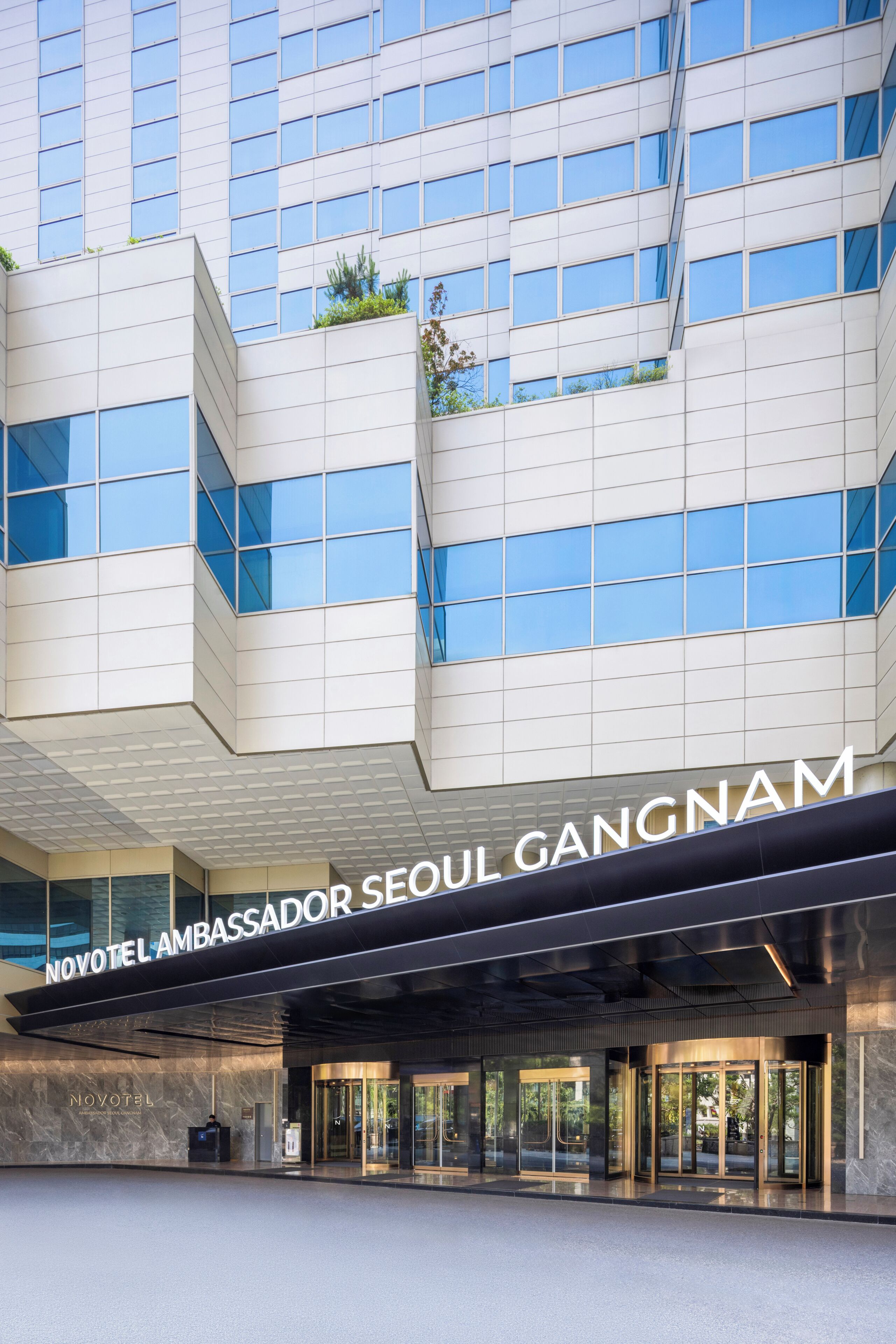 A modern hotel entrance with a glass facade, a reception desk, and the name 'NOVOTEL AMBASSADOR SEOUL GANGNAM' prominently displayed.