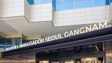 A modern hotel entrance with a glass facade, a reception desk, and the name 'NOVOTEL AMBASSADOR SEOUL GANGNAM' prominently displayed.