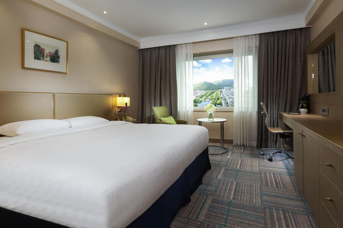 Une chambre d’hôtel avec un grand lit, un bureau, une chaise, une petite table avec un vase, et une vue sur le paysage urbain par la fenêtre.