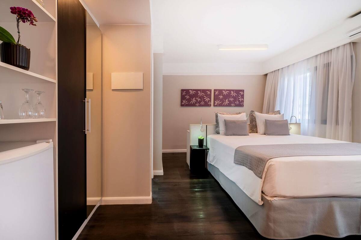 Una habitación de hotel con cama, mesitas de noche, una planta en maceta y una lámpara fijada a la pared.