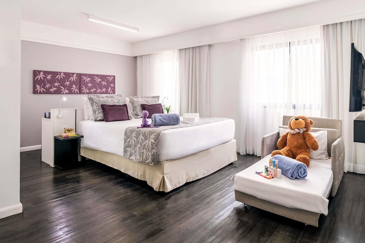 Quarto de hotel moderno com uma cama grande, um sofá, um urso de pelúcia e uma televisão.