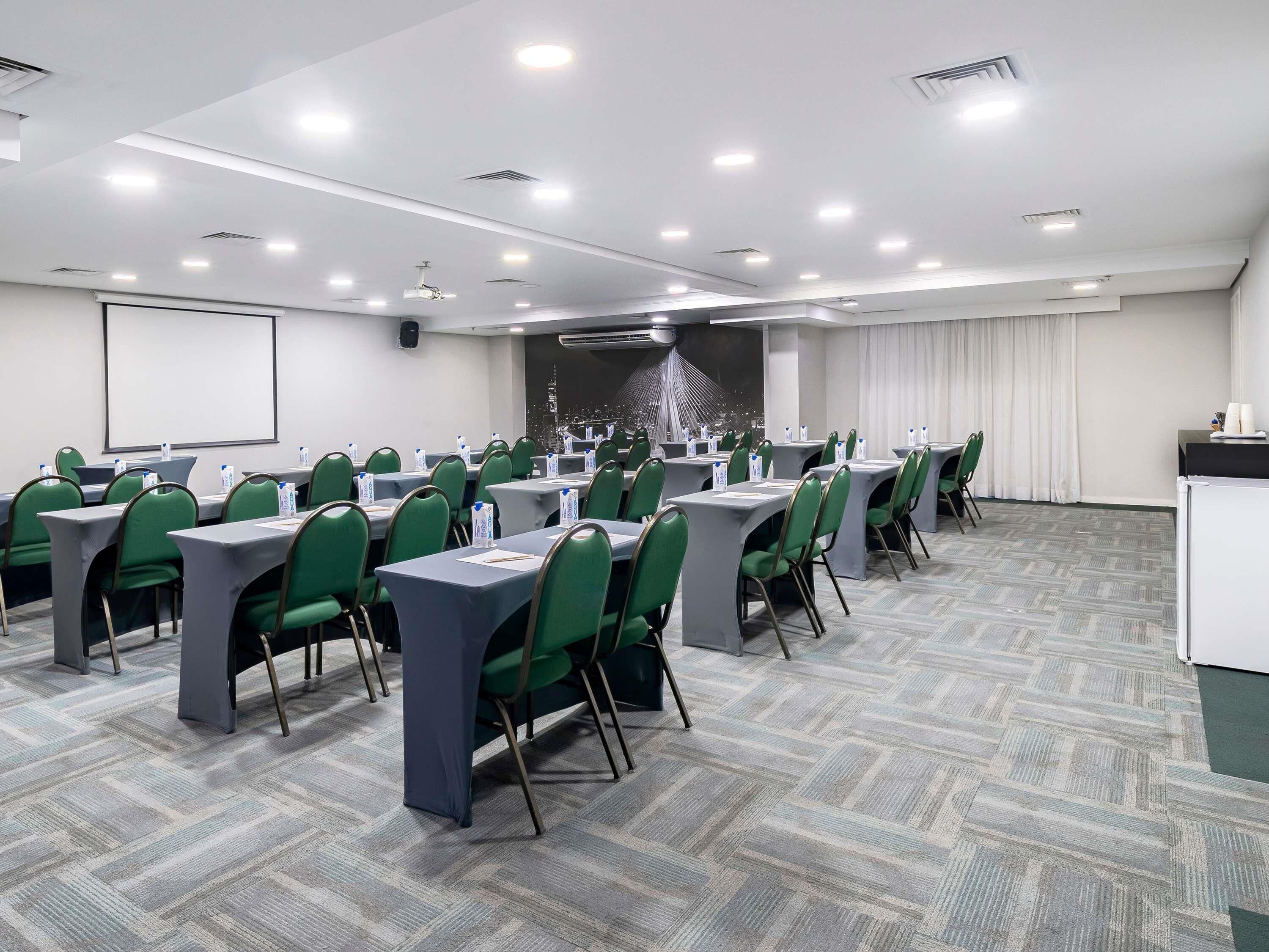 Ein Konferenzraum mit grünen Stühlen, Tischen, einer Projektionsleinwand und einer Stadtansicht als Wandbild.