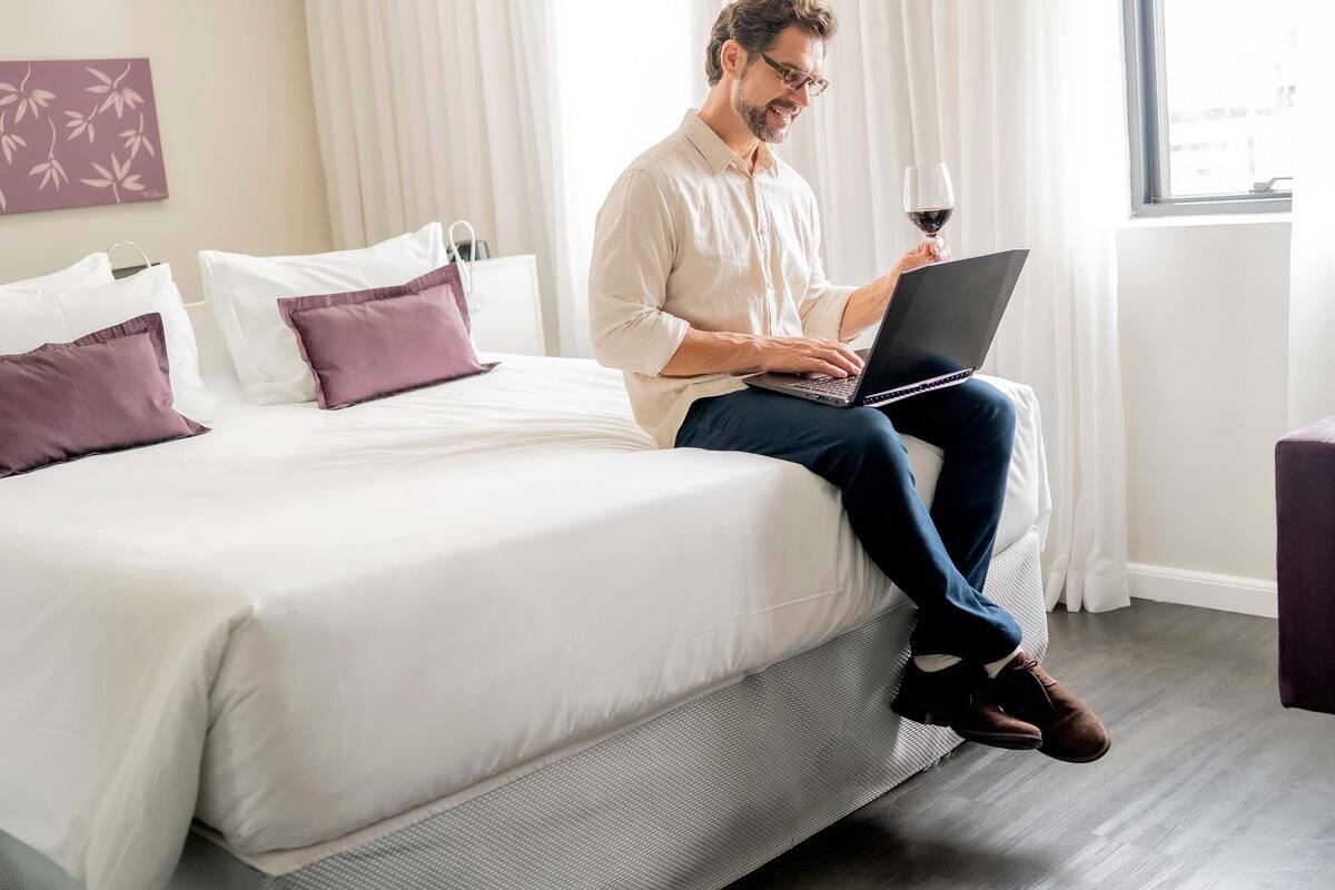 Una persona sentada en una cama con un ordenador portátil y una copa de vino, en una habitación con ventana y cortinas.