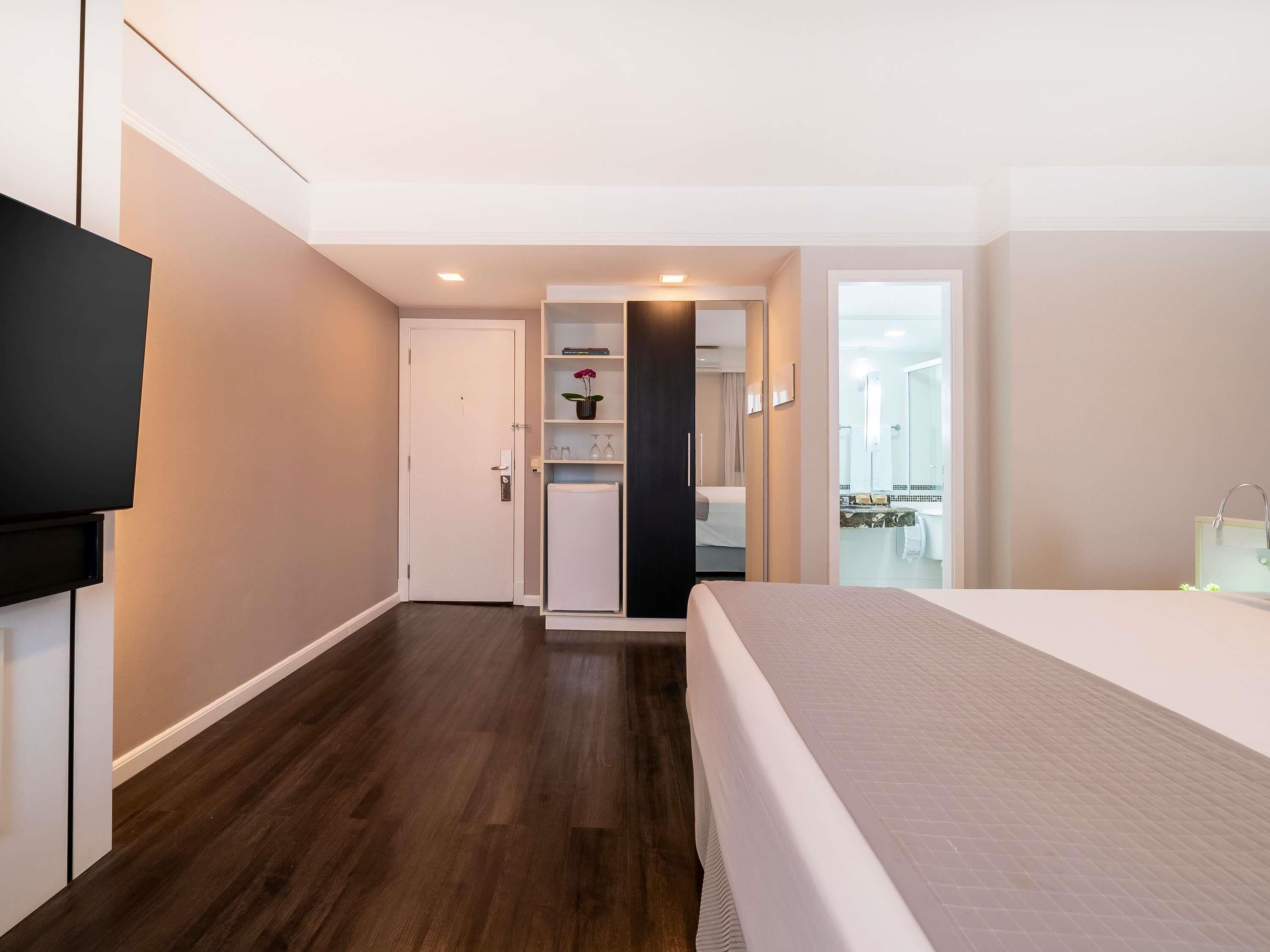 A hotel room with a bed, a TV, a door leading to a bathroom, and a small refrigerator.
