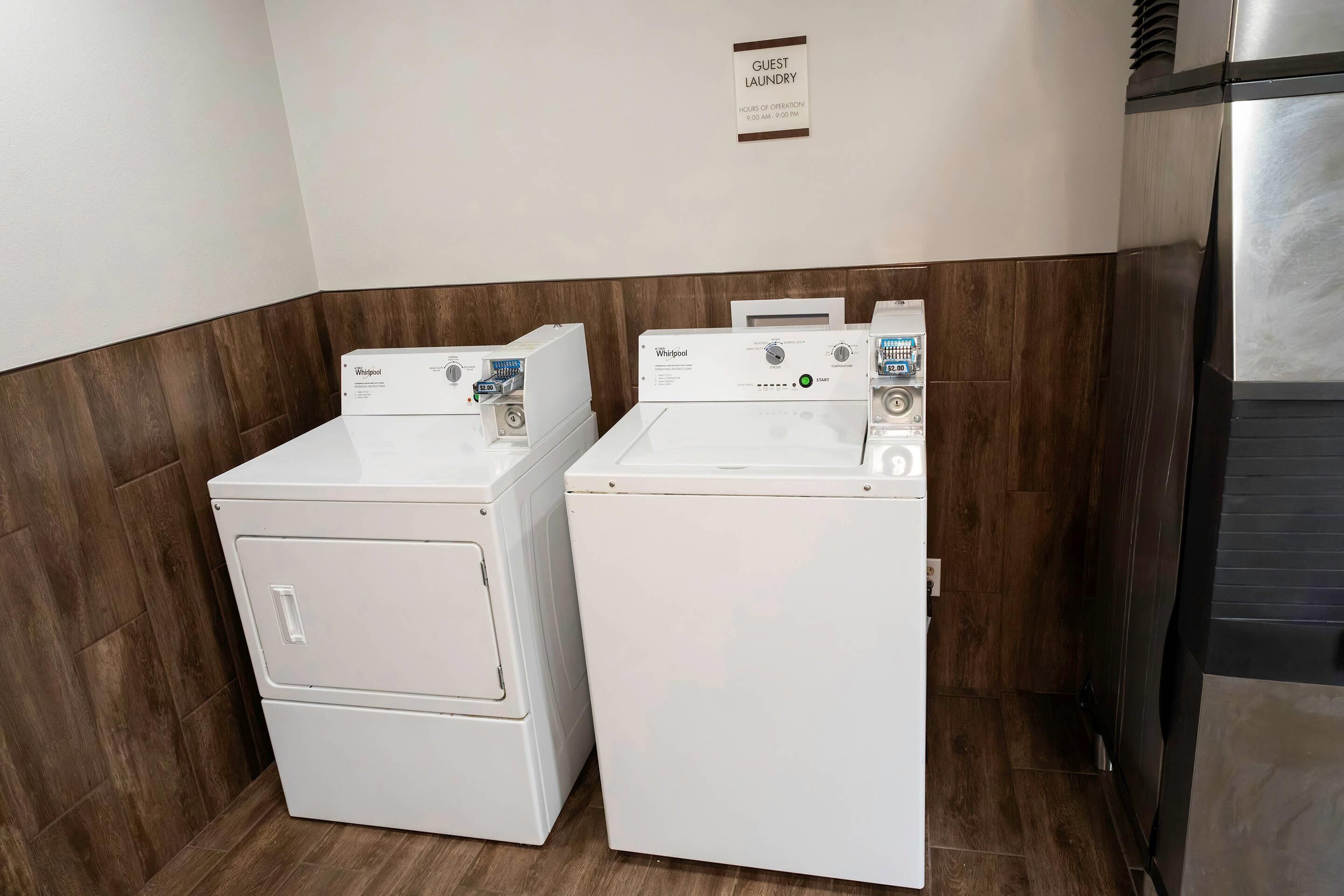 Uma área de lavanderia com duas máquinas de lavar e secar Whirlpool, uma placa com os horários de funcionamento da lavanderia para hóspedes e uma geladeira de aço inoxidável.