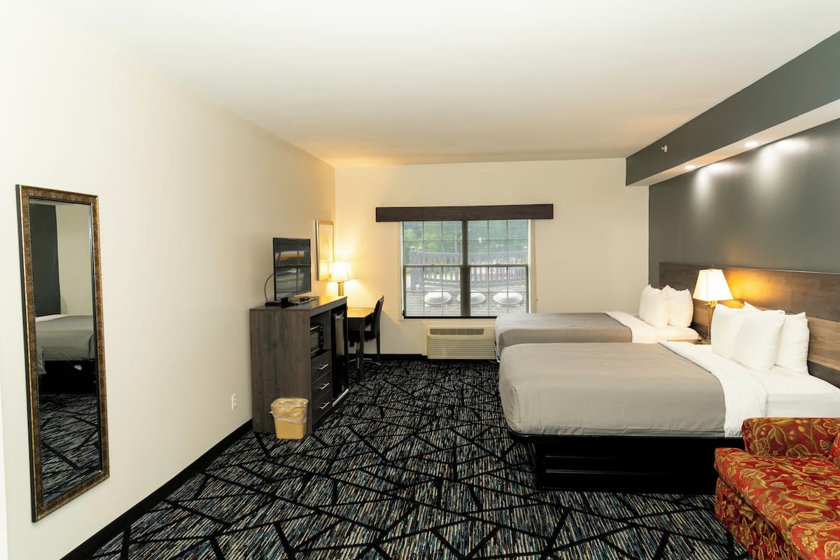 A hotel room with two beds, a desk with a chair, a dresser, a large mirror, and a window with a view of parked cars.