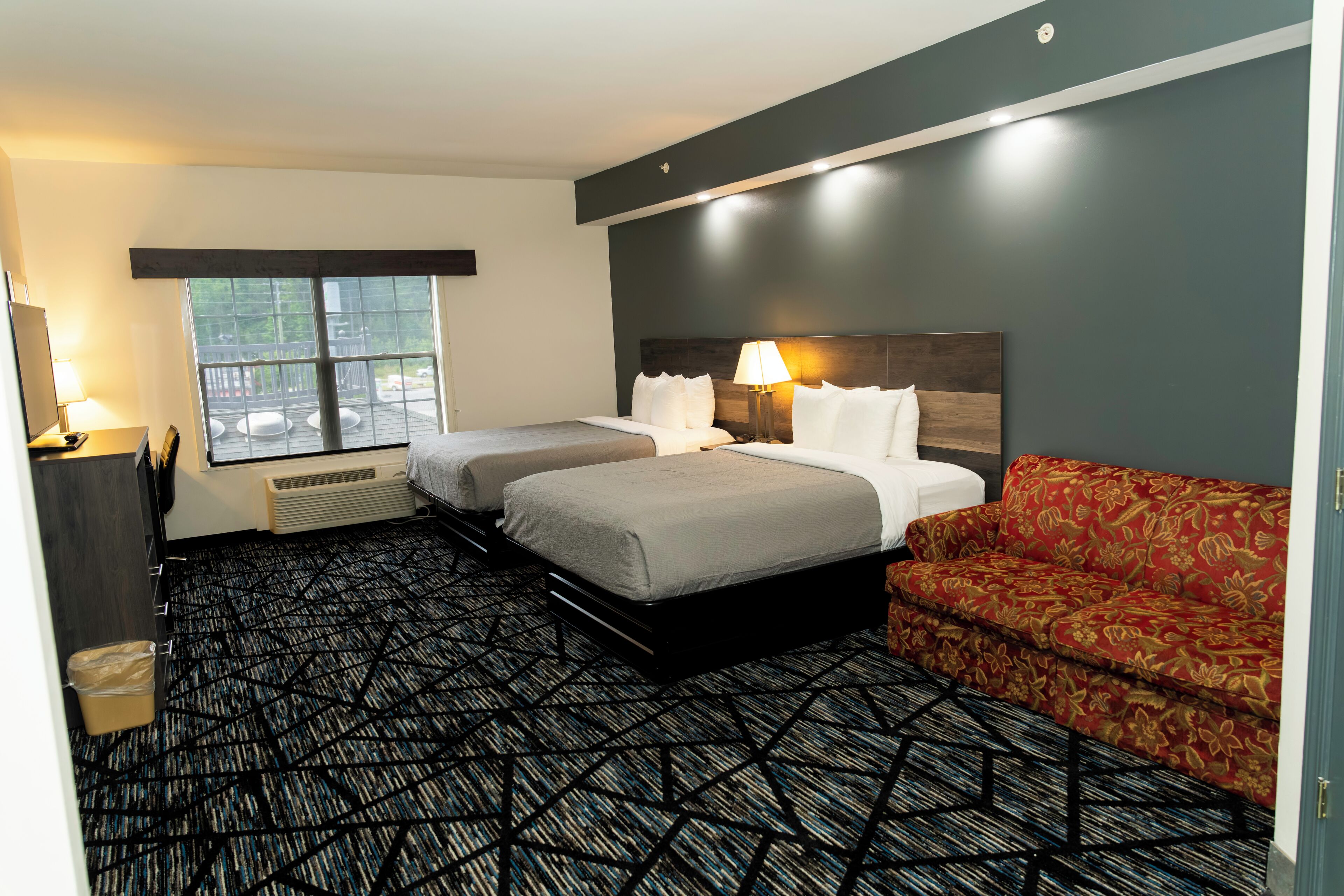 Una habitación de hotel con dos camas, un sofá rojo, un ventanal grande y una alfombra estampada.