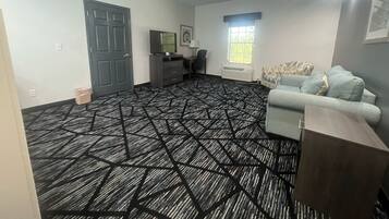 Una habitación con alfombra estampada, una puerta gris, un televisor sobre un mueble, una ventana y un sofá gris.