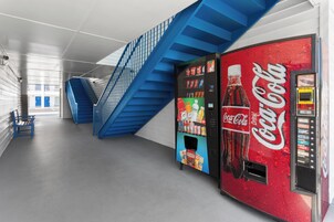 Una máquina expendedora de Coca-Cola en un edificio moderno, con una escalera azul y un banco.