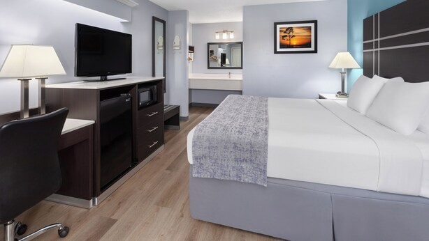 A modern hotel room with a flat-screen TV, a desk with a lamp, a bed with a blanket, a nightstand with a lamp, and a painting on the wall.