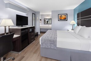 A modern hotel room with a flat-screen TV, a desk with a lamp, a bed with a blanket, a nightstand with a lamp, and a painting on the wall. - Baymont by Wyndham Brunswick GA (Brunswick)