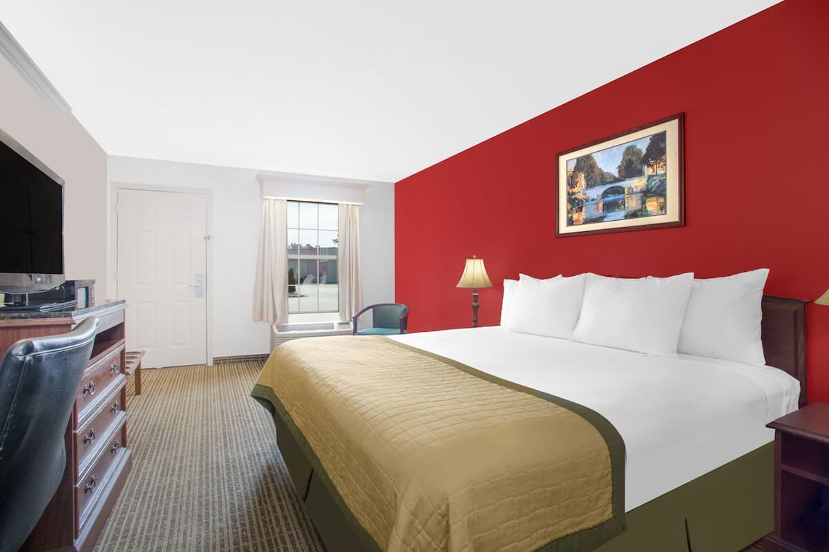A hotel room with a bed, a desk with a television, a chair, a window with curtains, and a painting on the wall.