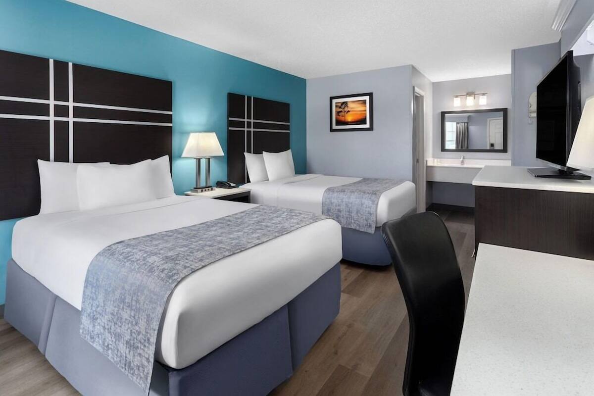 A hotel room with two beds, a desk, a chair, a TV, a lamp, a mirror, and a framed picture on the wall.