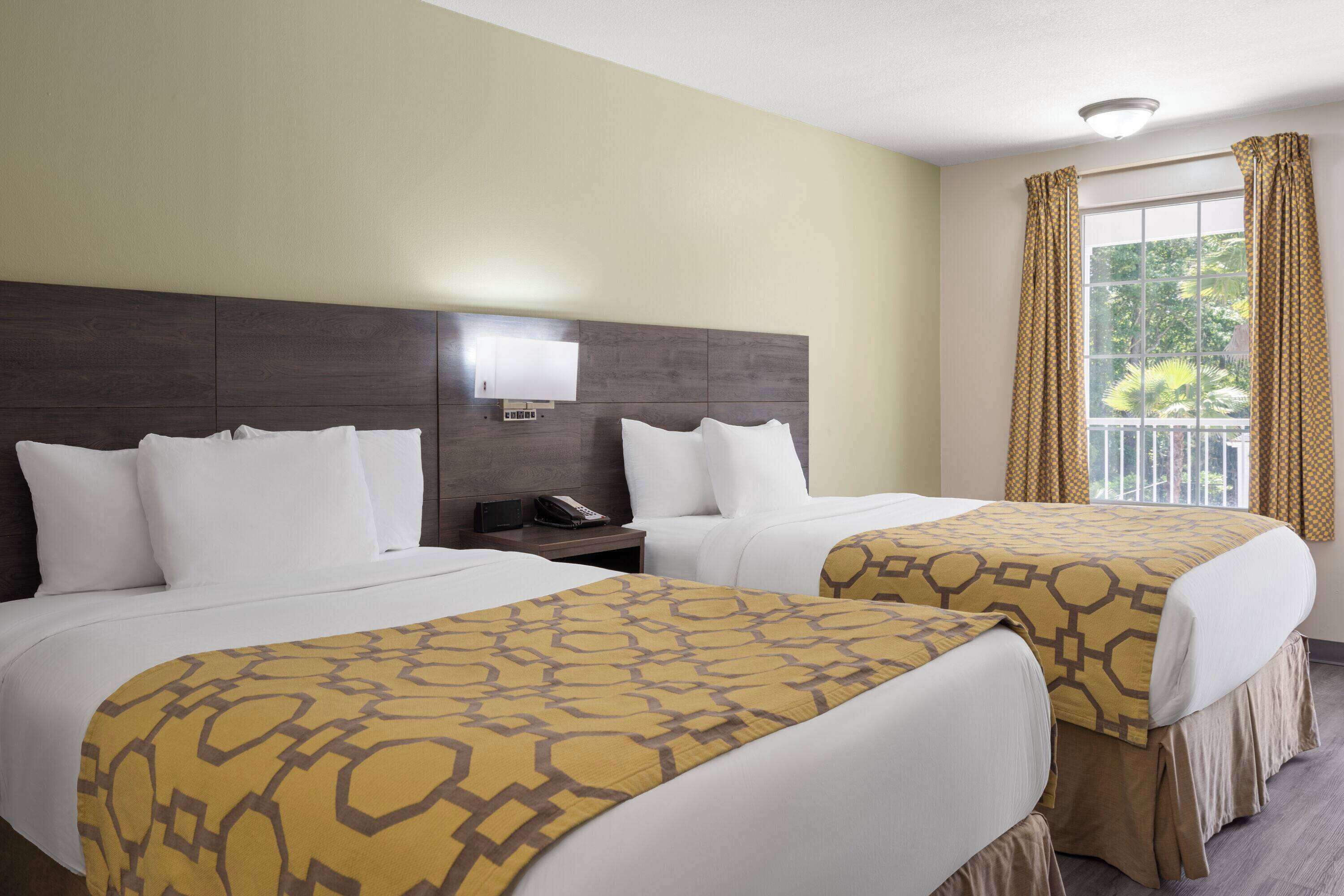 A hotel room with two beds, a bedside table with a phone, a wall-mounted light, and a window with curtains.