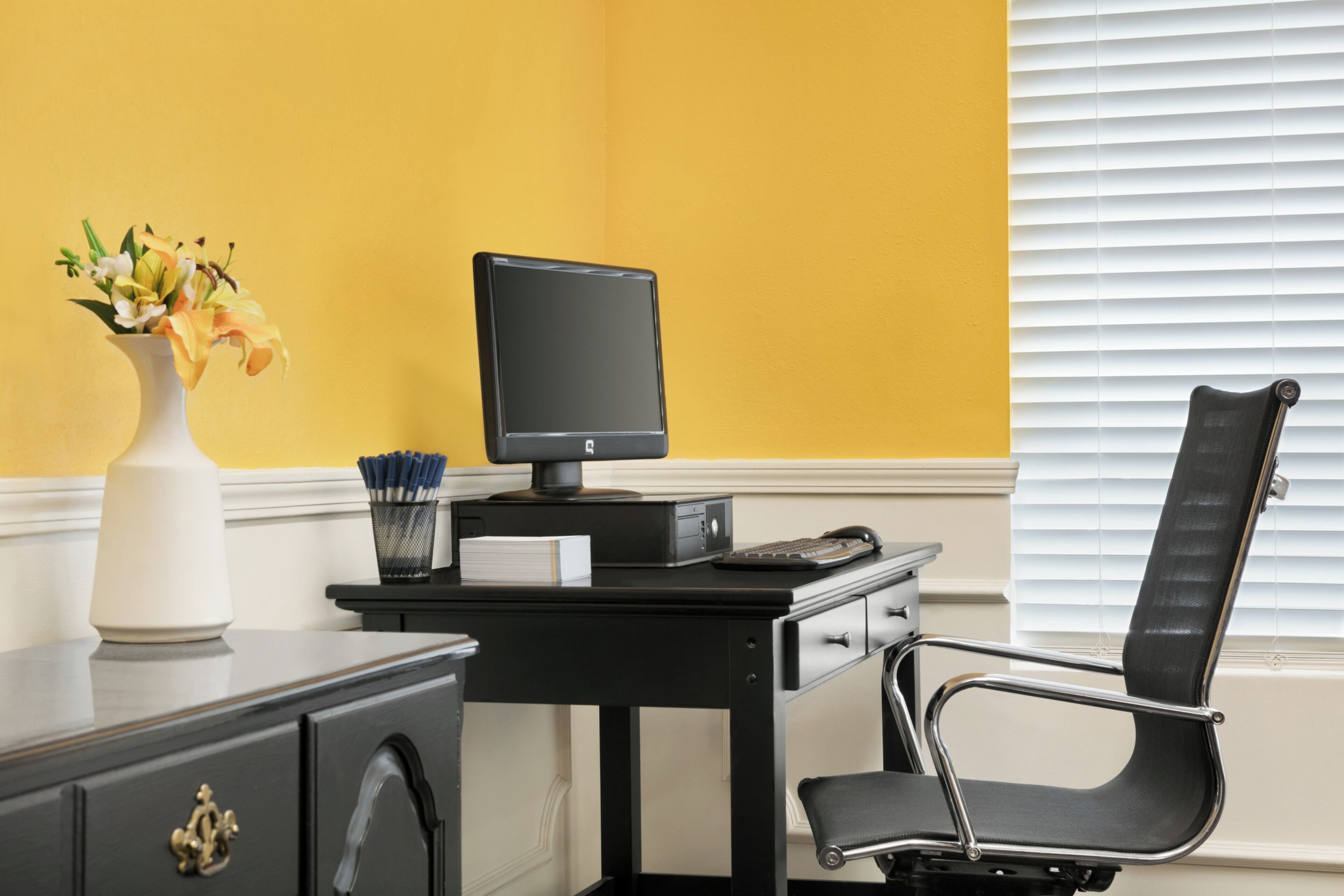 Ein schwarzer Schreibtisch mit einem Computer, einer Blumenvase und einem schwarzen Bürostuhl.