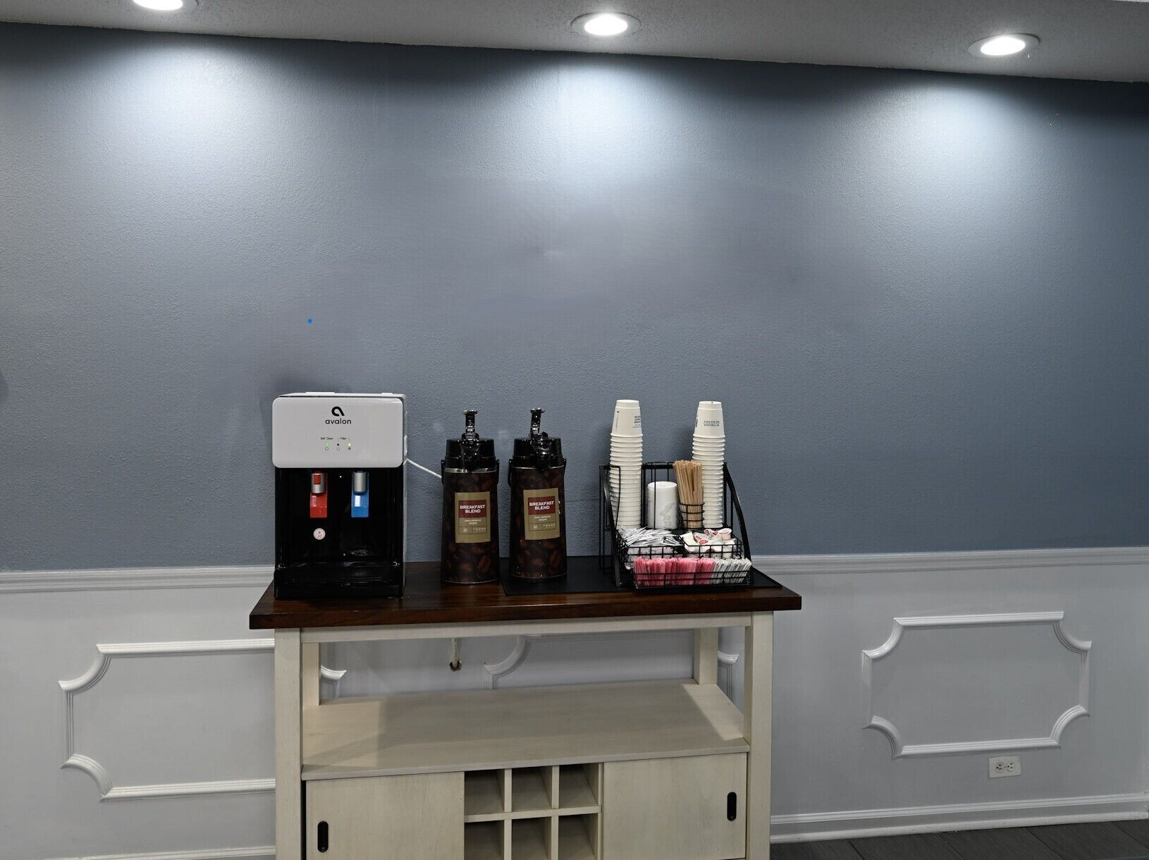 A coffee station with an espresso machine, two coffee dispensers, and a stack of disposable cups.