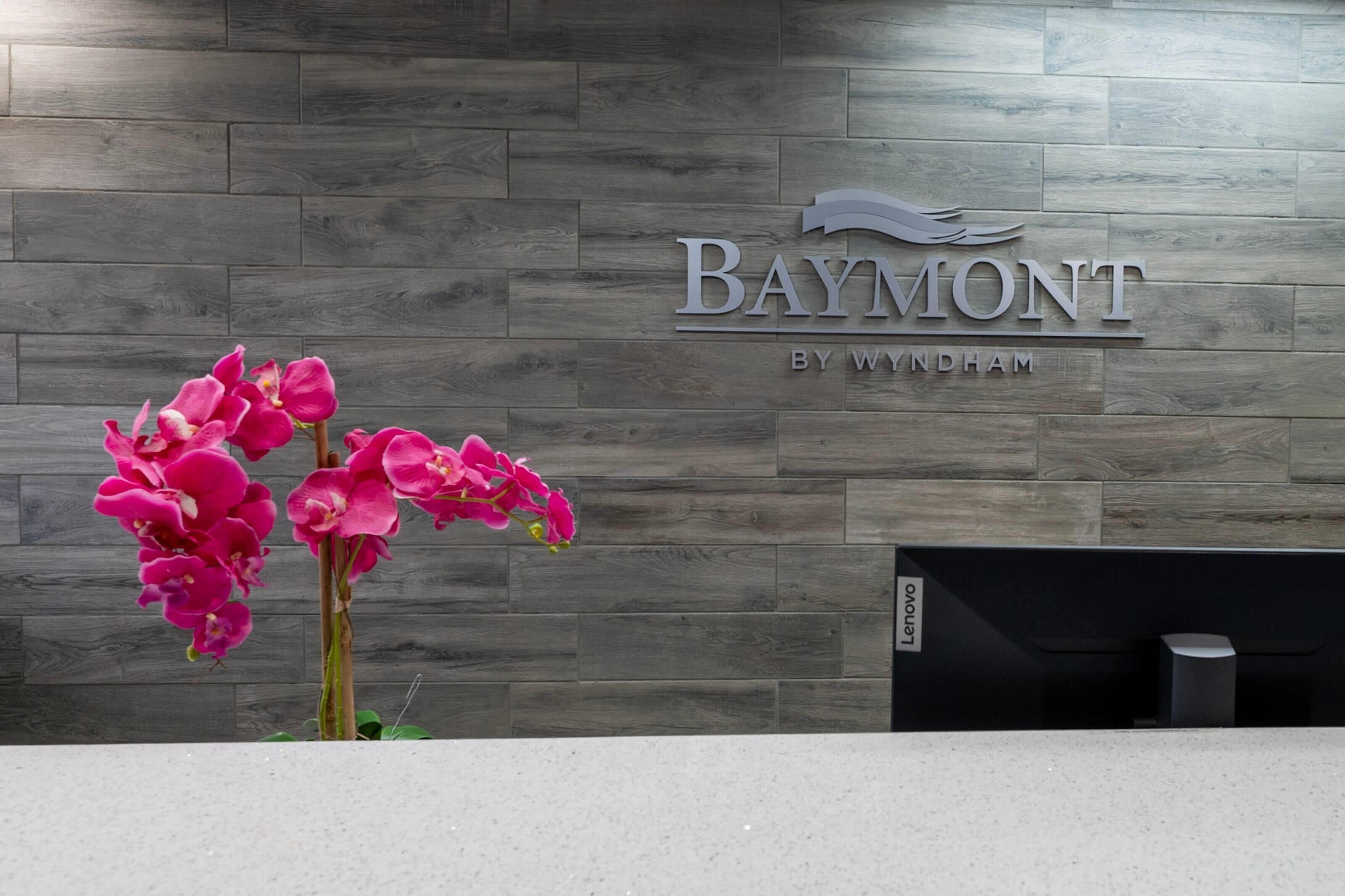 A reception desk with a wooden wall background, a sign that reads 'Baymont by Wyndham,' and a flower arrangement.