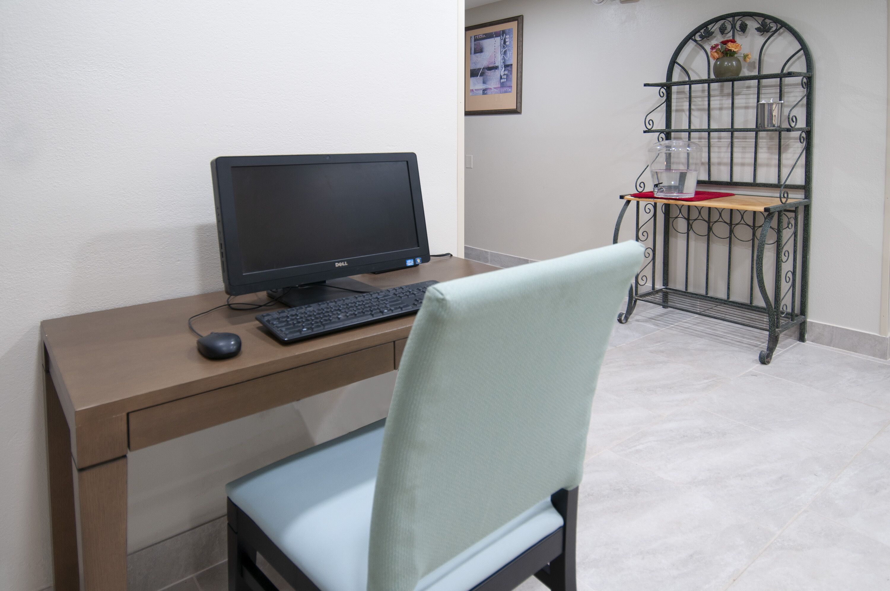 A computer desk with a monitor, keyboard, and mouse. There is a chair in front of the desk. A metal shelf with a vase and a picture frame is visible in the background.