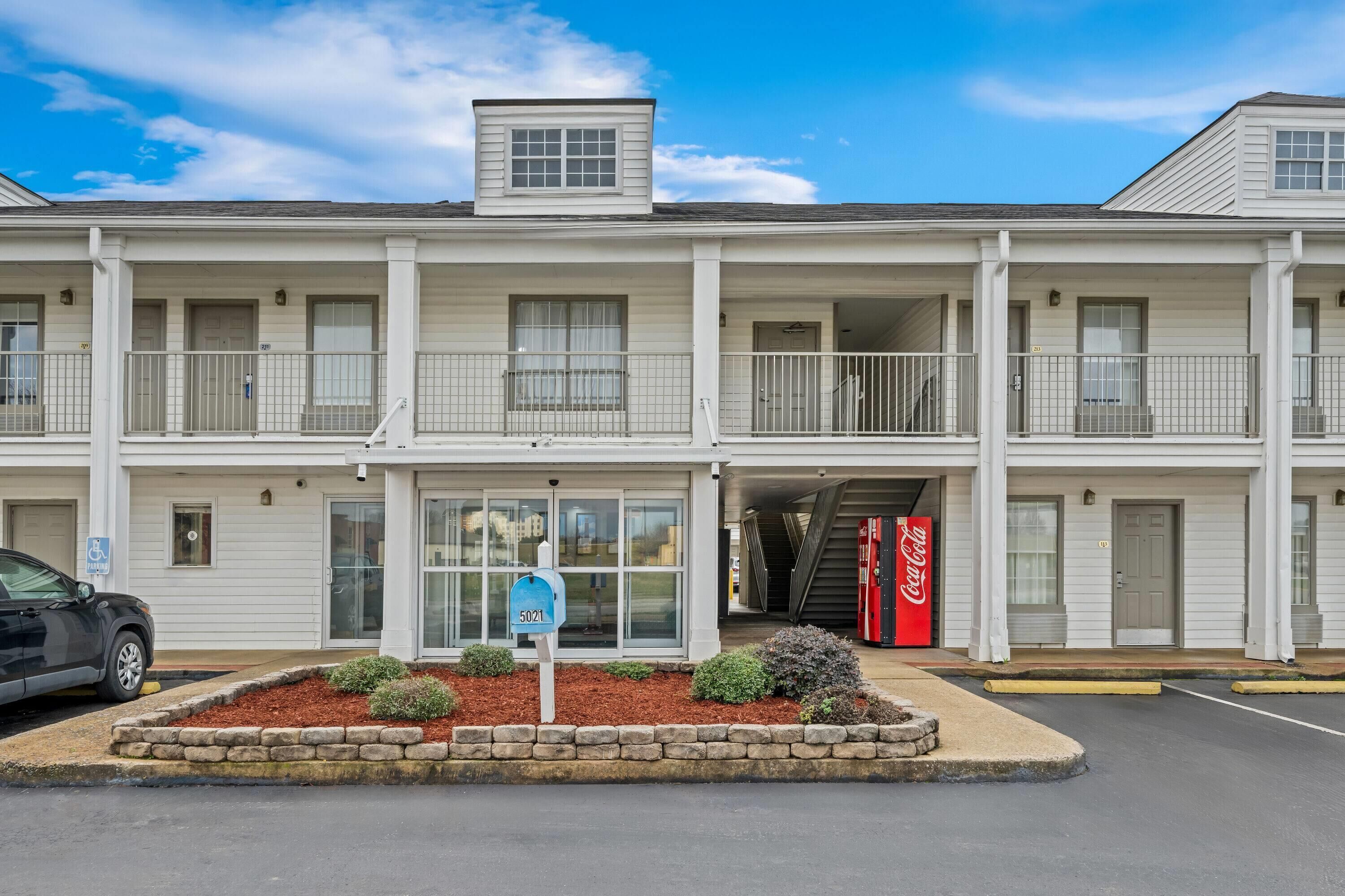 A multi-story motel with a central entrance, a Coca-Cola vending machine, and a parking sign.