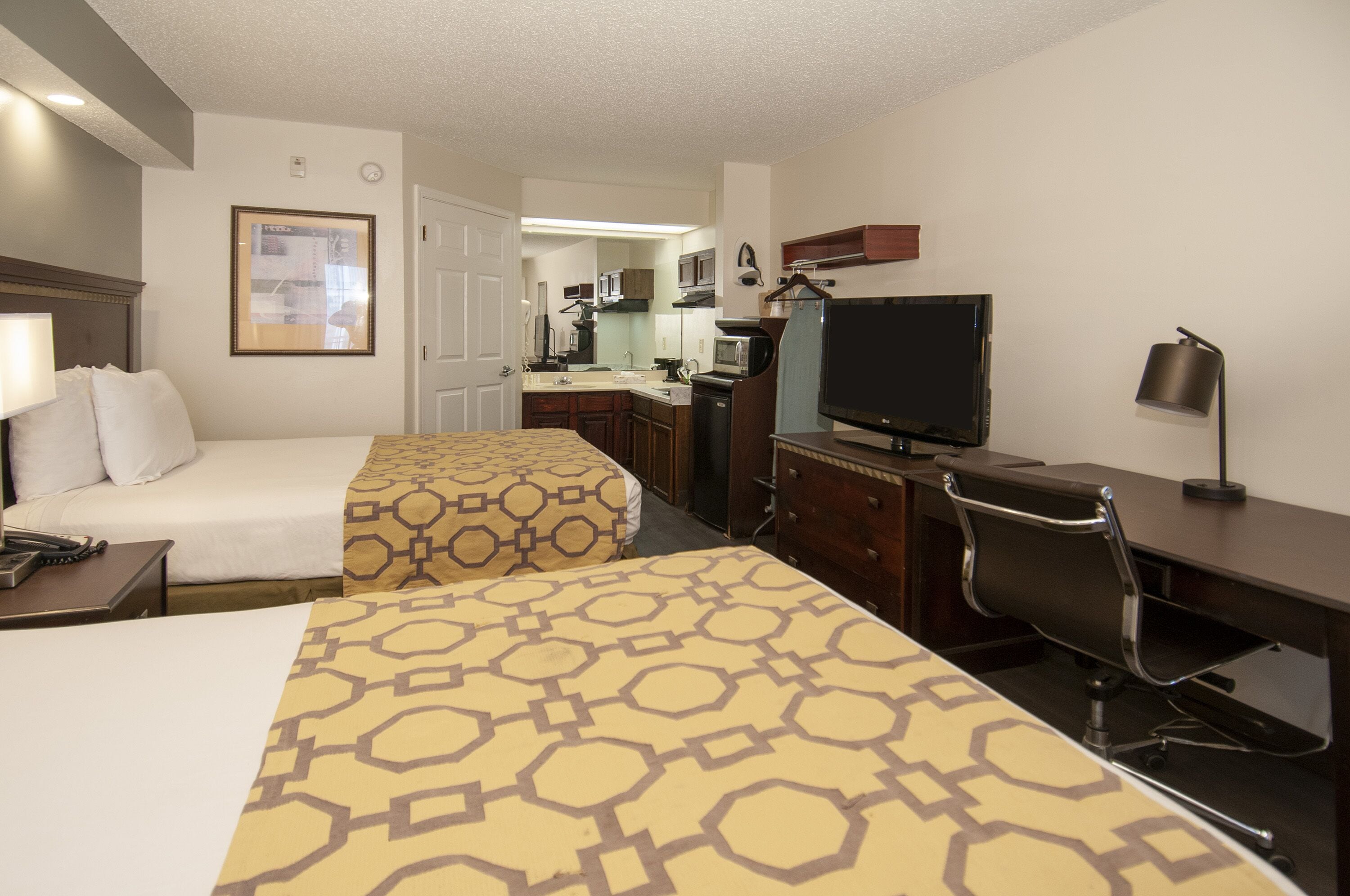 A hotel room with two beds, a desk, a TV, and a kitchenette.