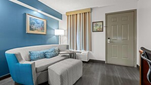 A modern hotel room with a blue accent wall, a gray sofa with a matching ottoman, a window with vertical blinds, a door, and a framed picture on the wall. - Baymont by Wyndham Tuscaloosa (Tuscaloosa)