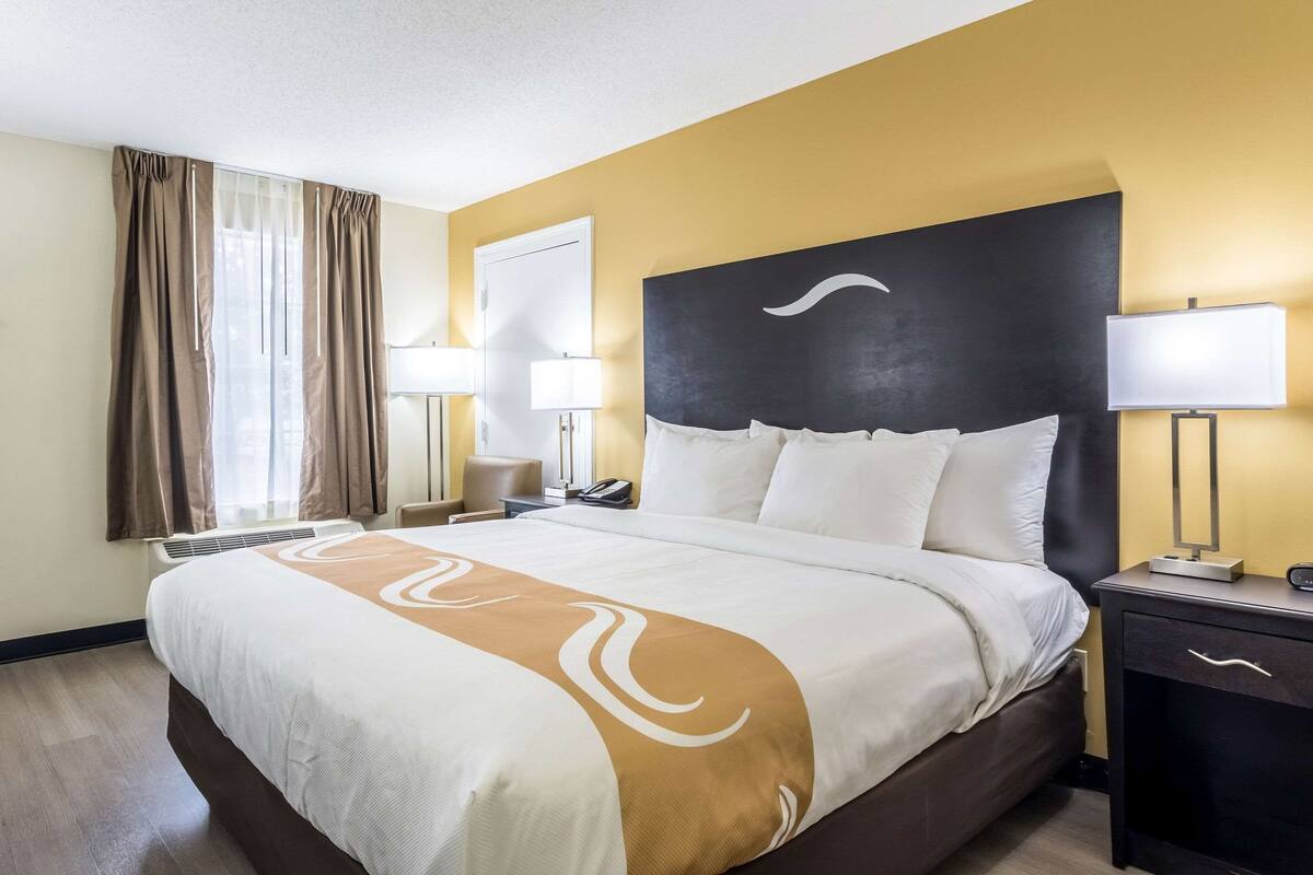 Quarto de hotel com uma cama grande, uma mesa de cabeceira com abajur, cortinas na janela.