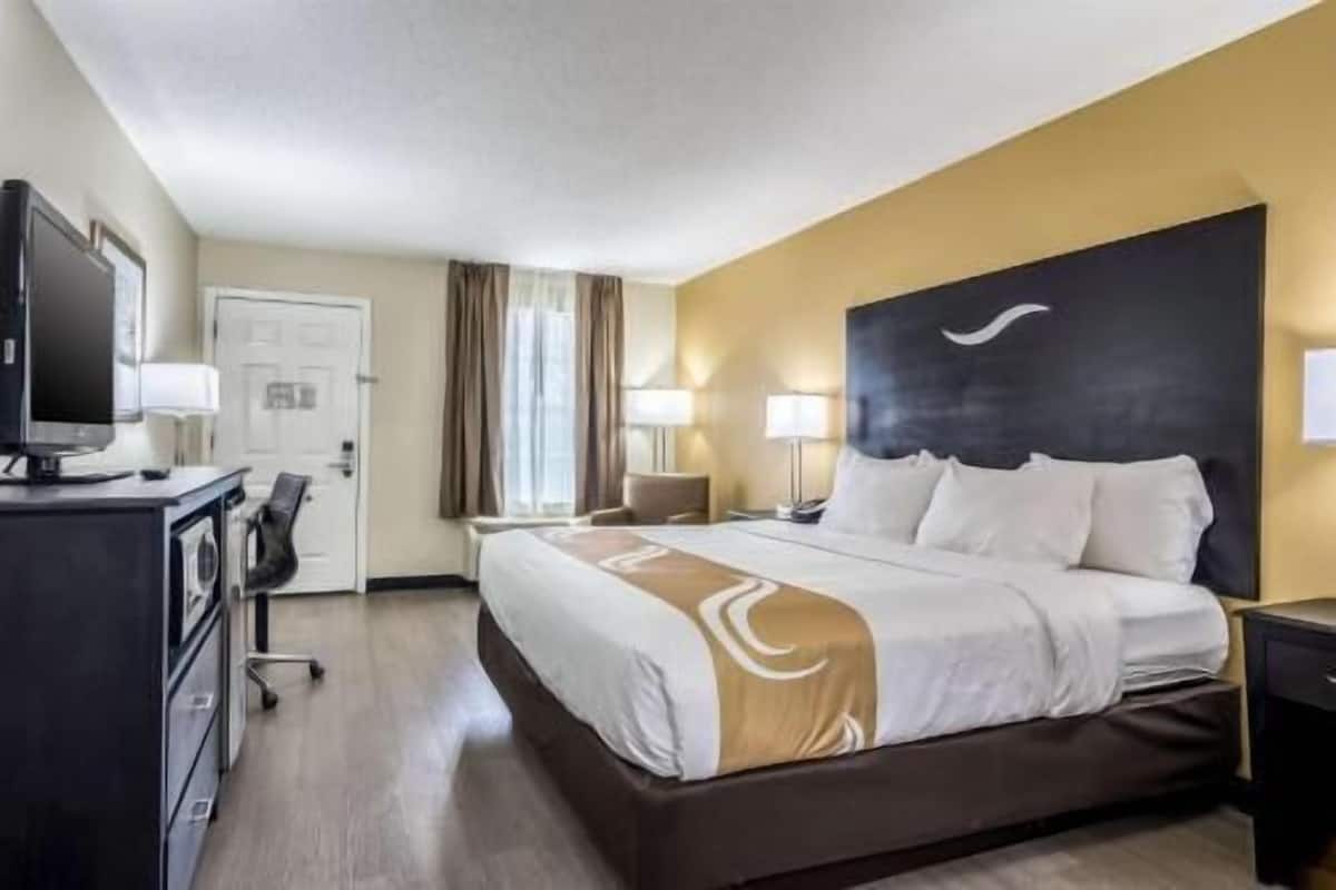 Quarto de hotel com cama, escrivaninha, cadeira, luminárias e televisão.