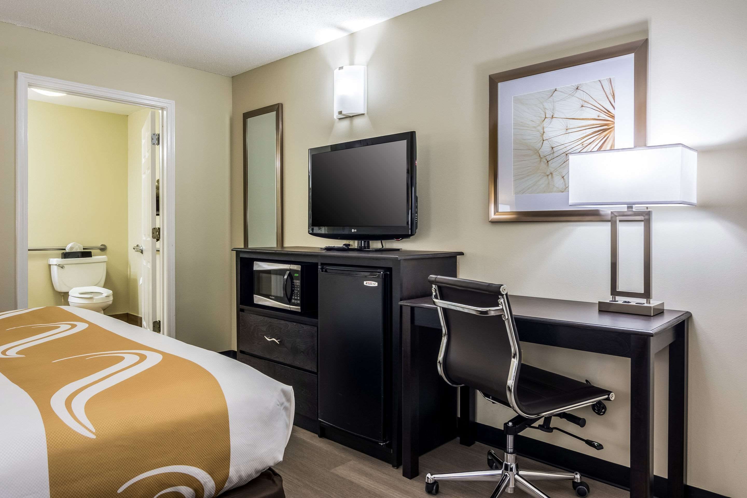 Una habitación de hotel con cama, escritorio con computadora, televisor, lámpara y una obra de arte enmarcada en la pared.