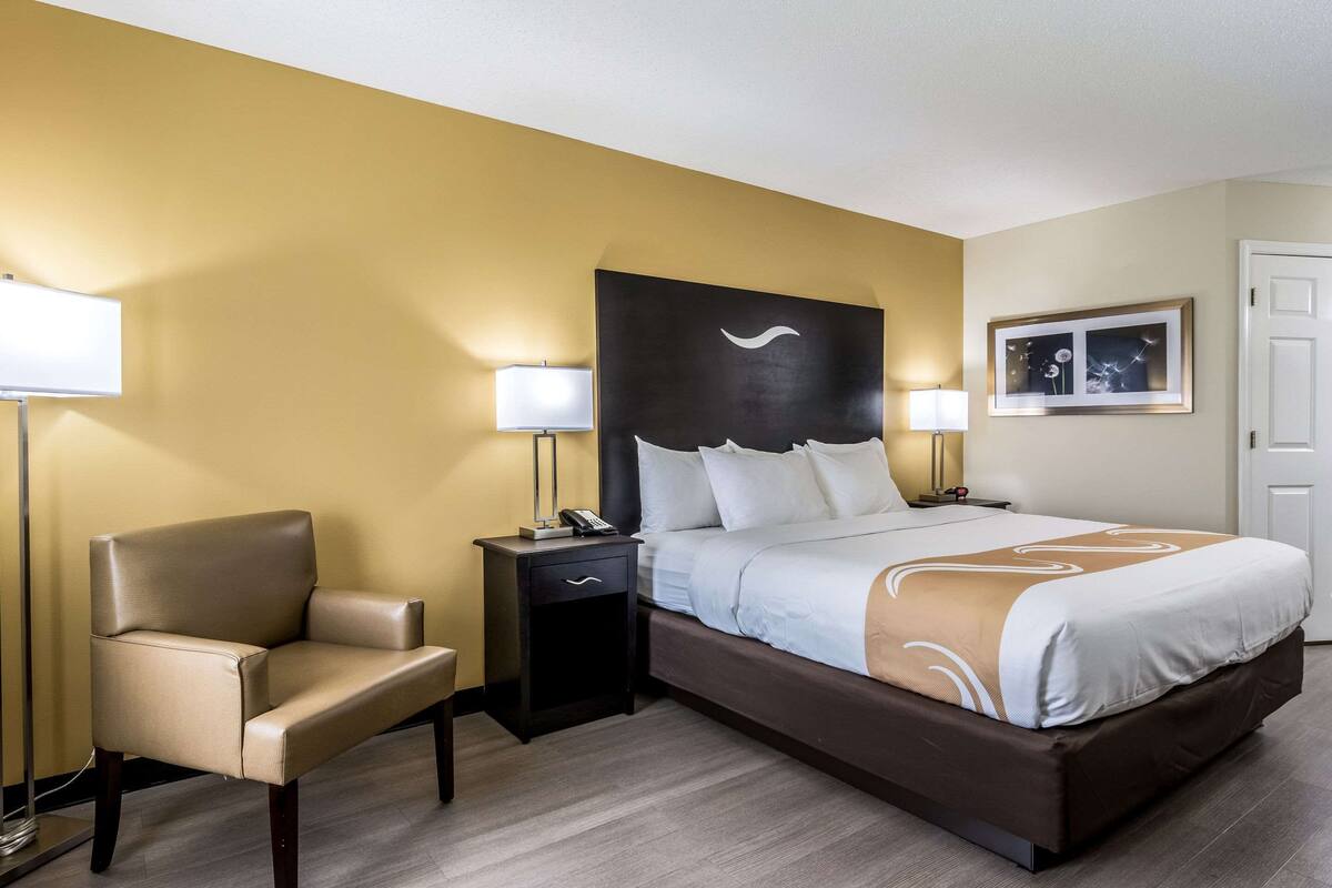 Quarto de hotel com uma cama, uma cadeira, mesa de cabeceira com telefone e luminária.
