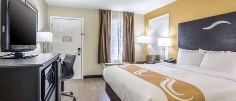 Een hotelkamer met een bed, een bureau met een computer, een stoel, een televisie en een raam met gordijnen.