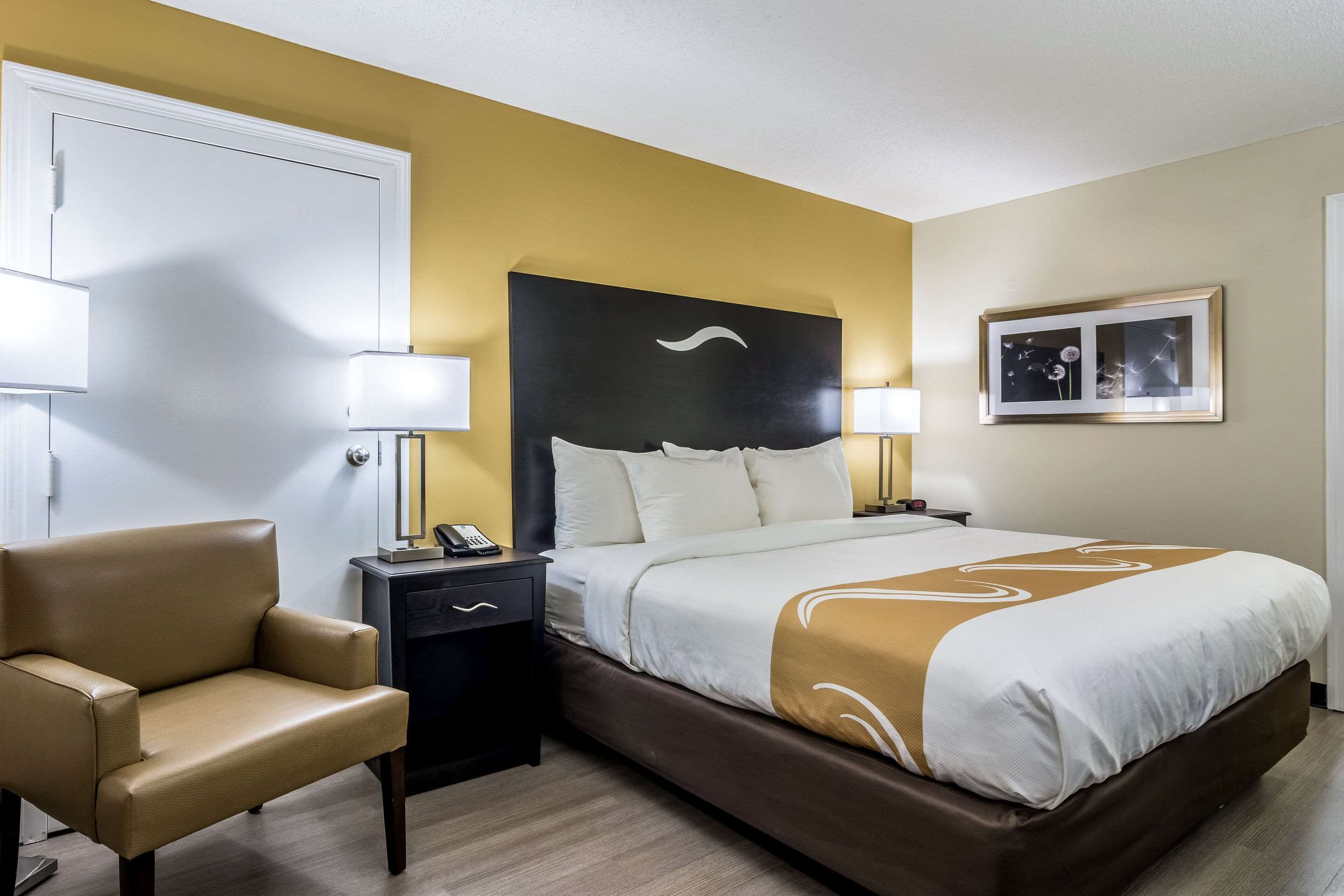 A hotel room with a bed, a chair, a nightstand, and a wall-mounted picture.