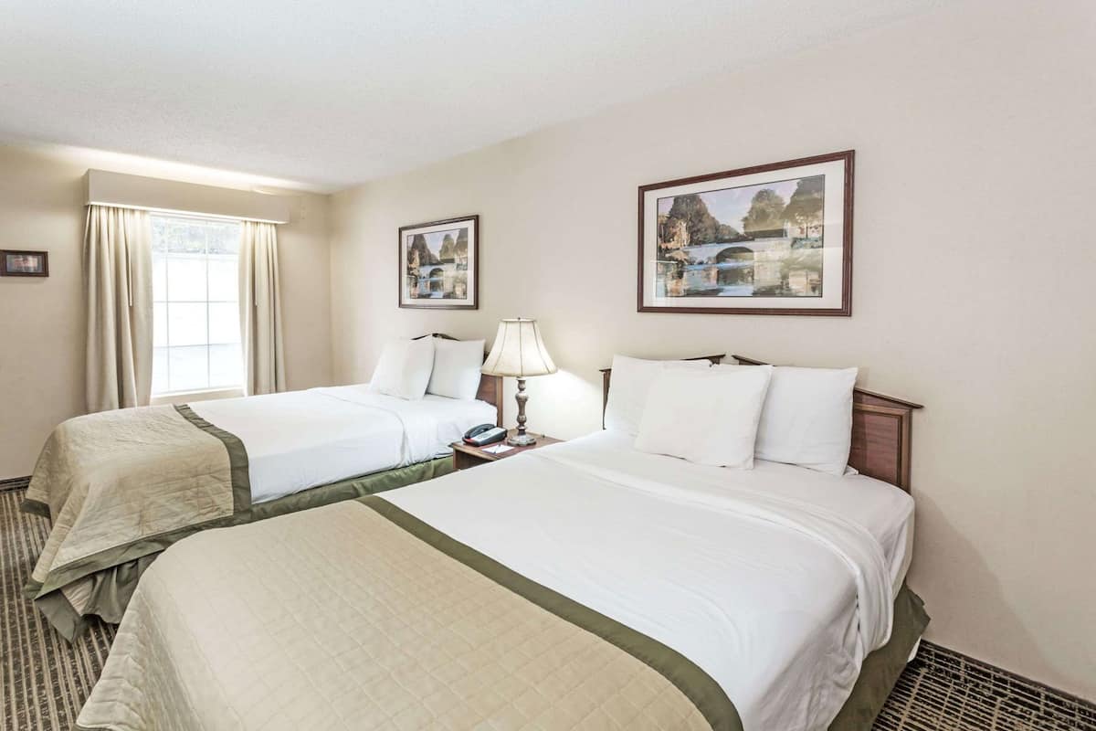 A hotel room with two beds, a window with curtains, a bedside table with a lamp, and two framed pictures on the wall.