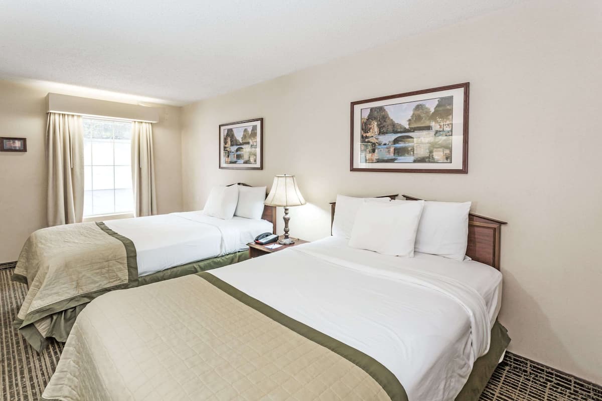 A hotel room with two beds, a window with curtains, a bedside table with a lamp, and two framed pictures on the wall.