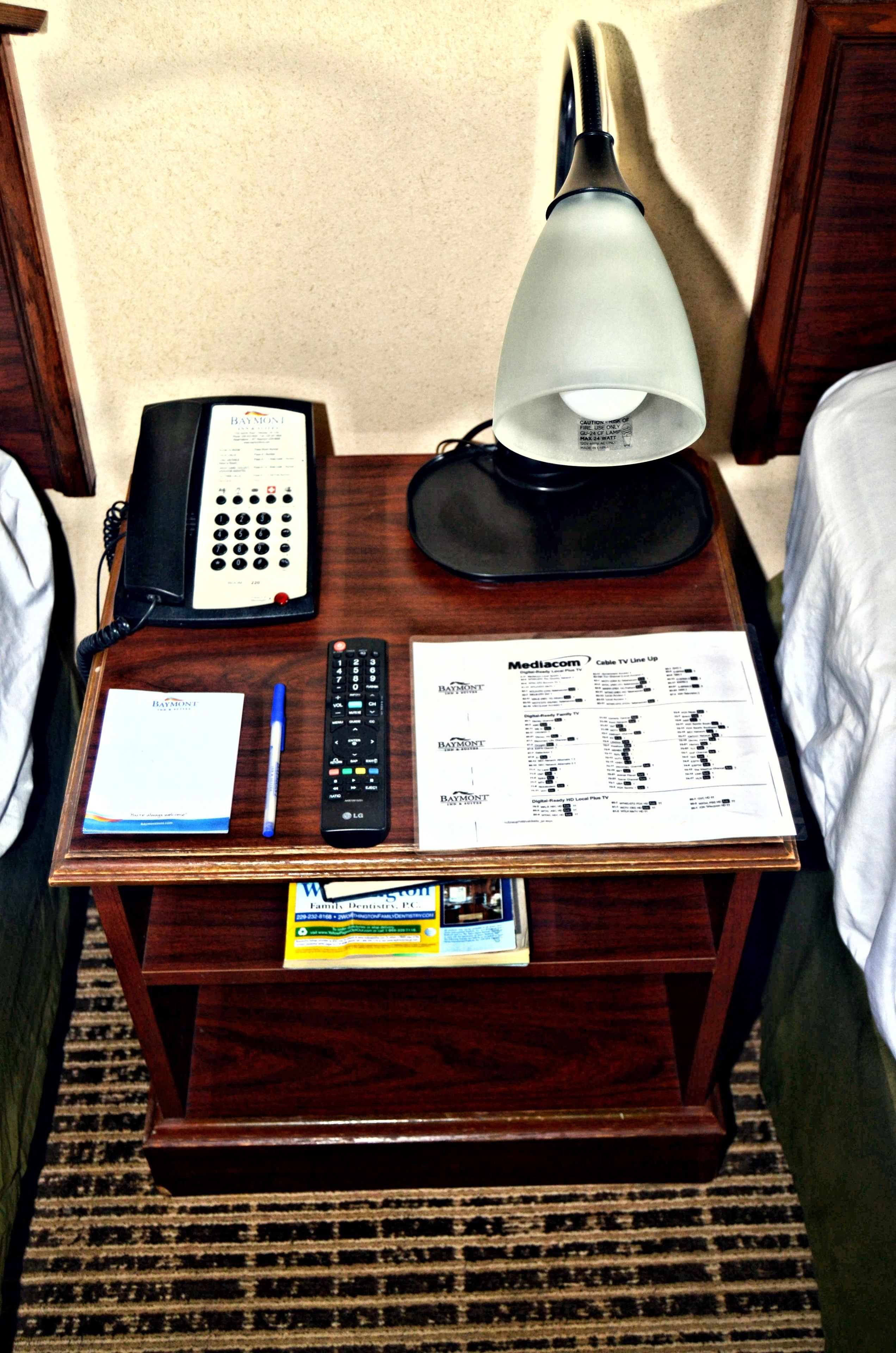 Une table de chevet en bois avec une lampe, un téléphone, une télécommande, un bloc-notes et un guide des programmes de la télévision par câble.