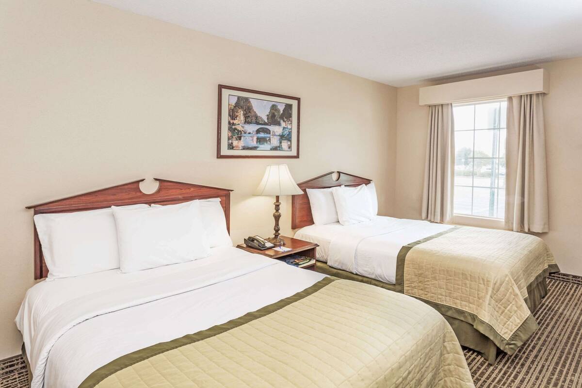 A hotel room with two beds, a nightstand with a lamp, a framed picture on the wall, and a window with curtains.