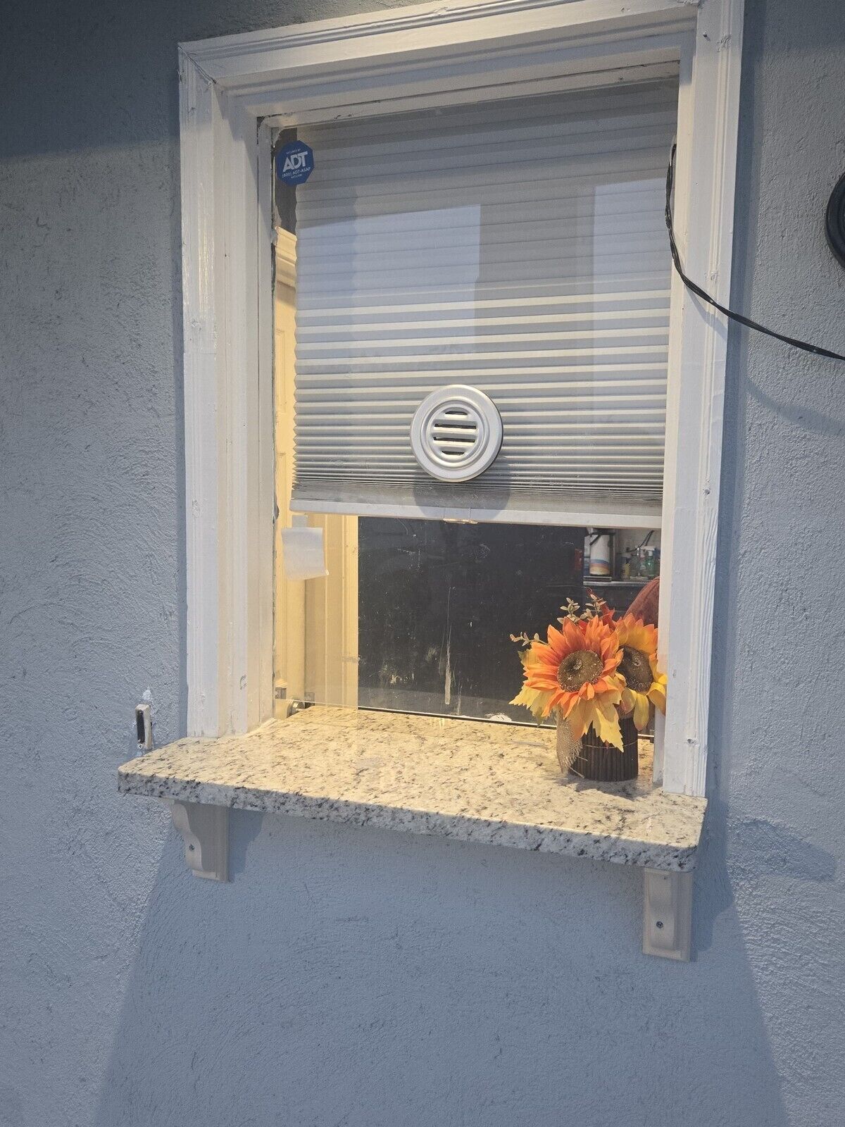 A window with a metal grill, a small shelf with a vase of flowers, and a blue sign above the window.