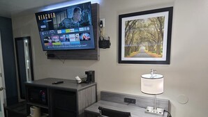 A hotel room with a TV displaying a streaming service interface, a desk with a lamp, a framed picture of a forest path, and a microwave. - Baymont by Wyndham Valdosta at Valdosta Mall (Valdosta)