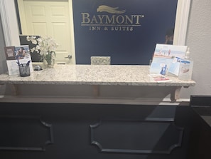 A reception desk with a Baymont Inn & Suites sign, a framed photo, a vase with flowers, a hand sanitizer dispenser, and informational brochures. - Baymont by Wyndham Valdosta at Valdosta Mall (Valdosta)