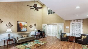 A spacious living room with a high ceiling, wooden flooring, a ceiling fan, a sofa, a coffee table, a desk with a chair, and a patterned rug. - Baymont by Wyndham Valdosta at Valdosta Mall (Valdosta)
