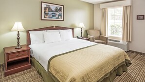 A hotel room with a large bed, two bedside tables with lamps, a chair, a window with curtains, and a painting on the wall. - Baymont by Wyndham Valdosta at Valdosta Mall (Valdosta)