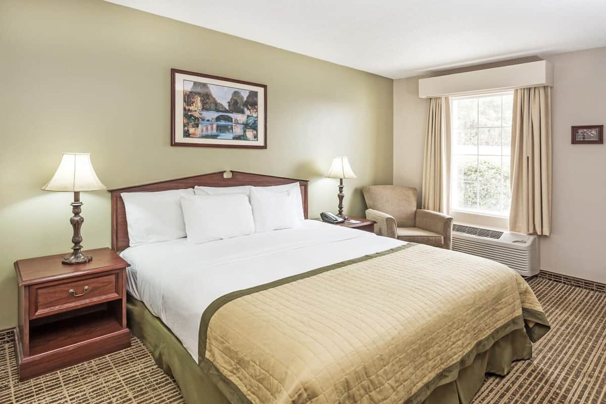 A hotel room with a bed, bedside lamps, a chair, a window with curtains, and a painting on the wall.