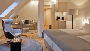 Et moderne soveværelse med en stor seng, et siddeområde med sofa og lænestol, et lille bord med lampe og et tekøkken.
