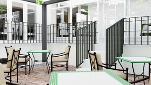 En udendørs patio med borde og stole, en pergola med grønt, samt udsigt til indretningen gennem store vinduer.
