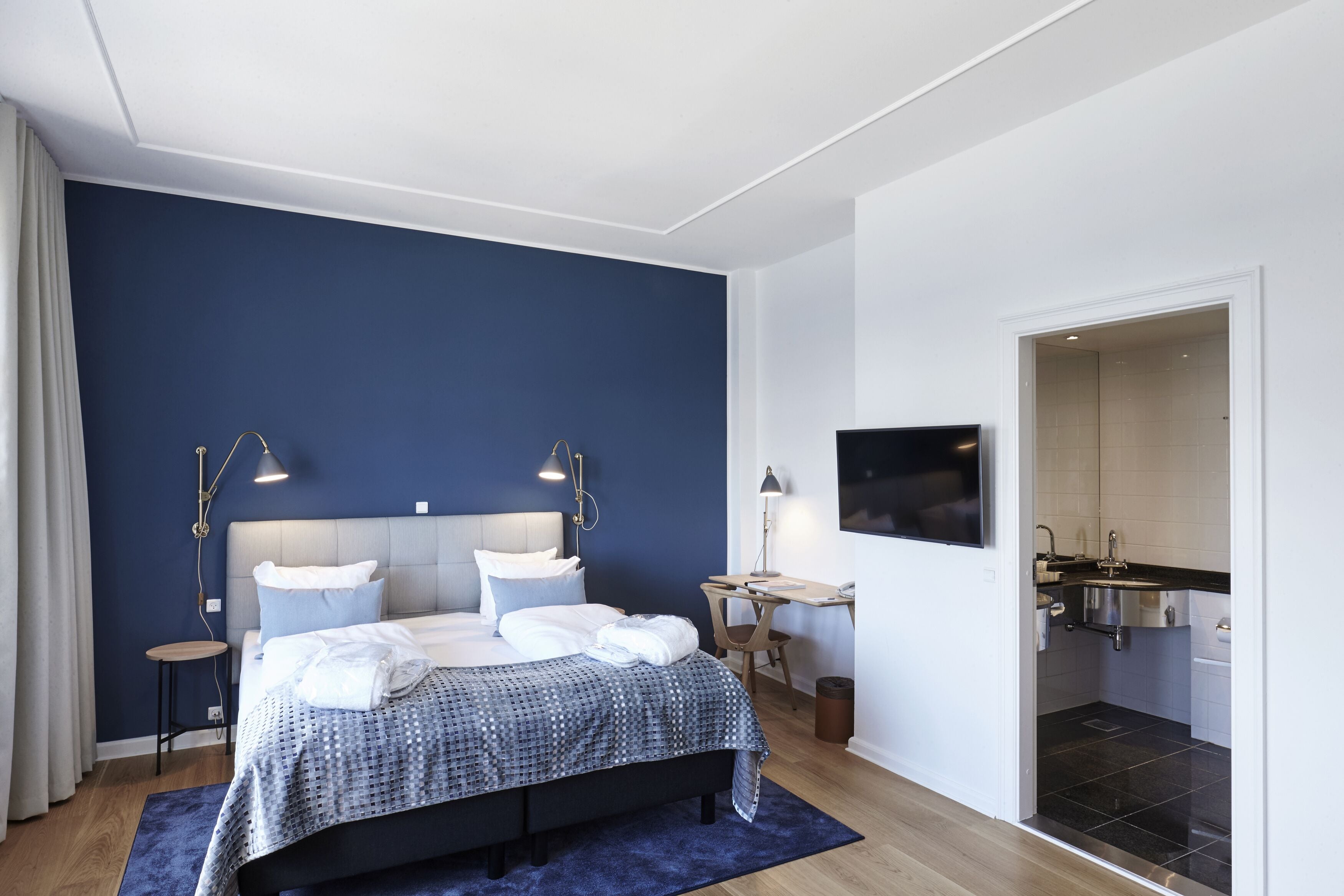 A modern bedroom with a large bed, bedside lamps, a desk, and a view into the bathroom.