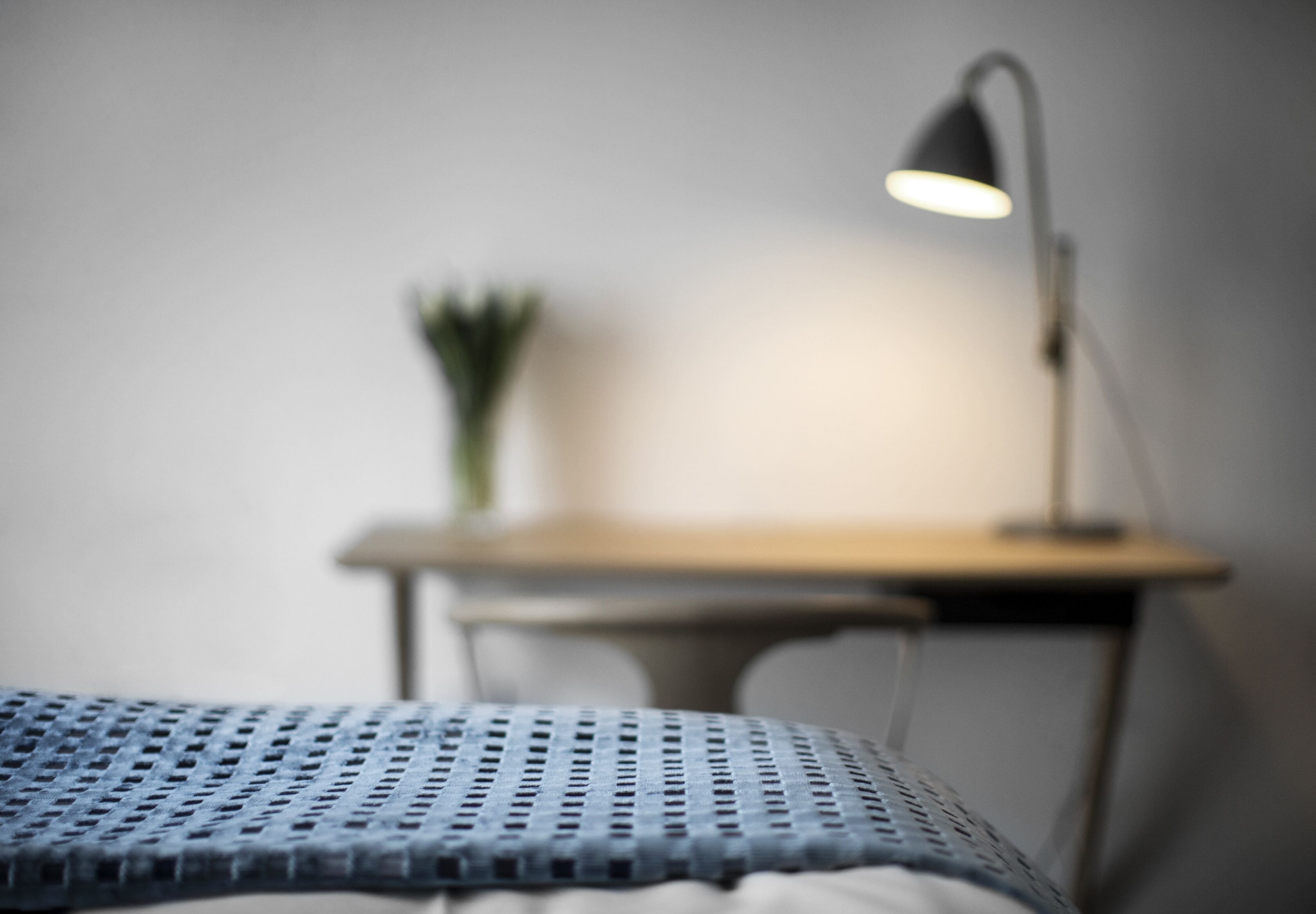 A bed with a blue textured cover, a bedside lamp, and a small table with a plant in the background.
