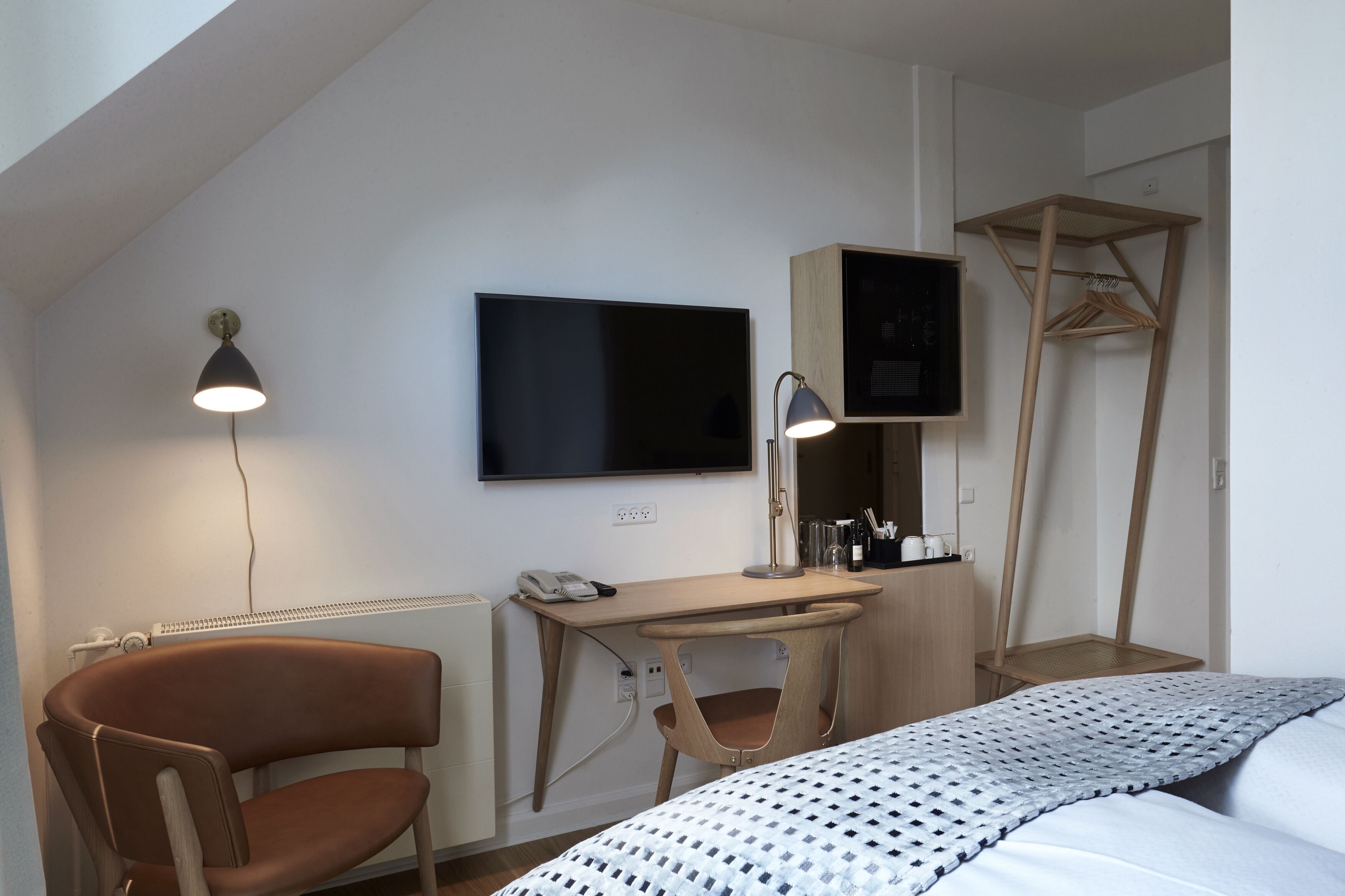 A modern hotel room with a bed, a desk with a computer, a chair, a TV mounted on the wall, and a hanging clothes rack.