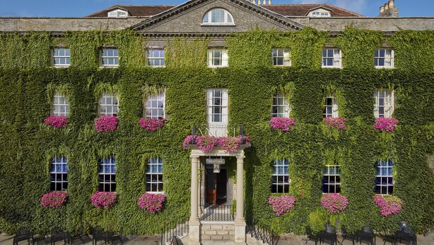 A historic building with a facade covered in ivy and pink flowers, featuring a central entrance with a sign reading "WELCOME".