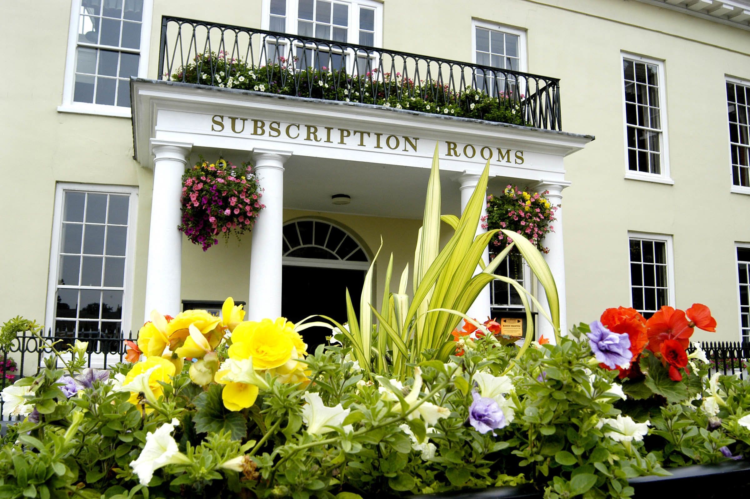 A building with a sign reading "SUBSCRIPTION ROOMS" above the entrance, surrounded by a variety of flowers.
