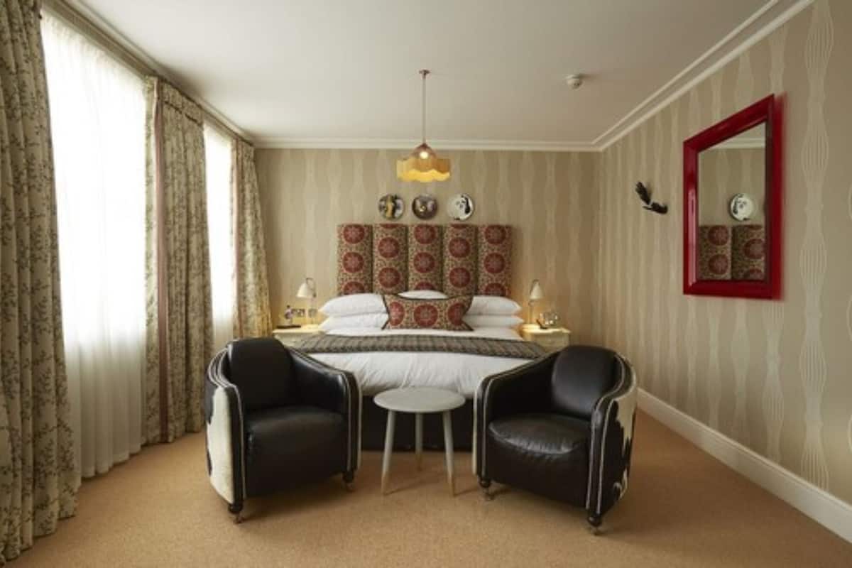 A hotel room with a bed, two chairs, a small table, a mirror, and a decorative headboard.