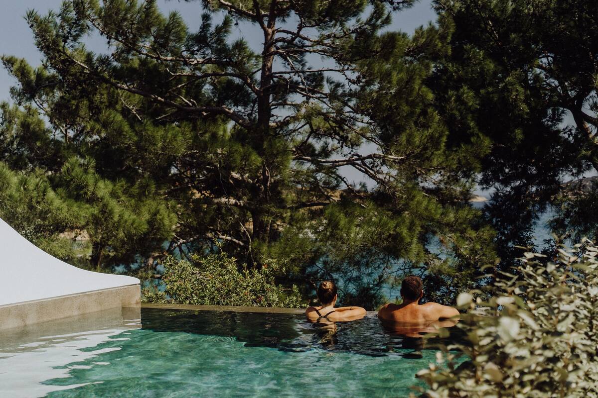 Piscina con dos personas, rodeada de árboles y una estructura blanca.